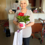 Older woman holds a pink potted flower and a white gift bag for "I Remember Mama" at Cobblestone Place apartments.
