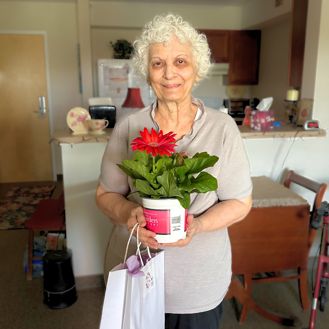 Older woman with white hair is smiling while holding a potted pink flower.