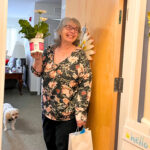Older Woman with gray hair smiles at her door as she holds a gift bag and flowers.