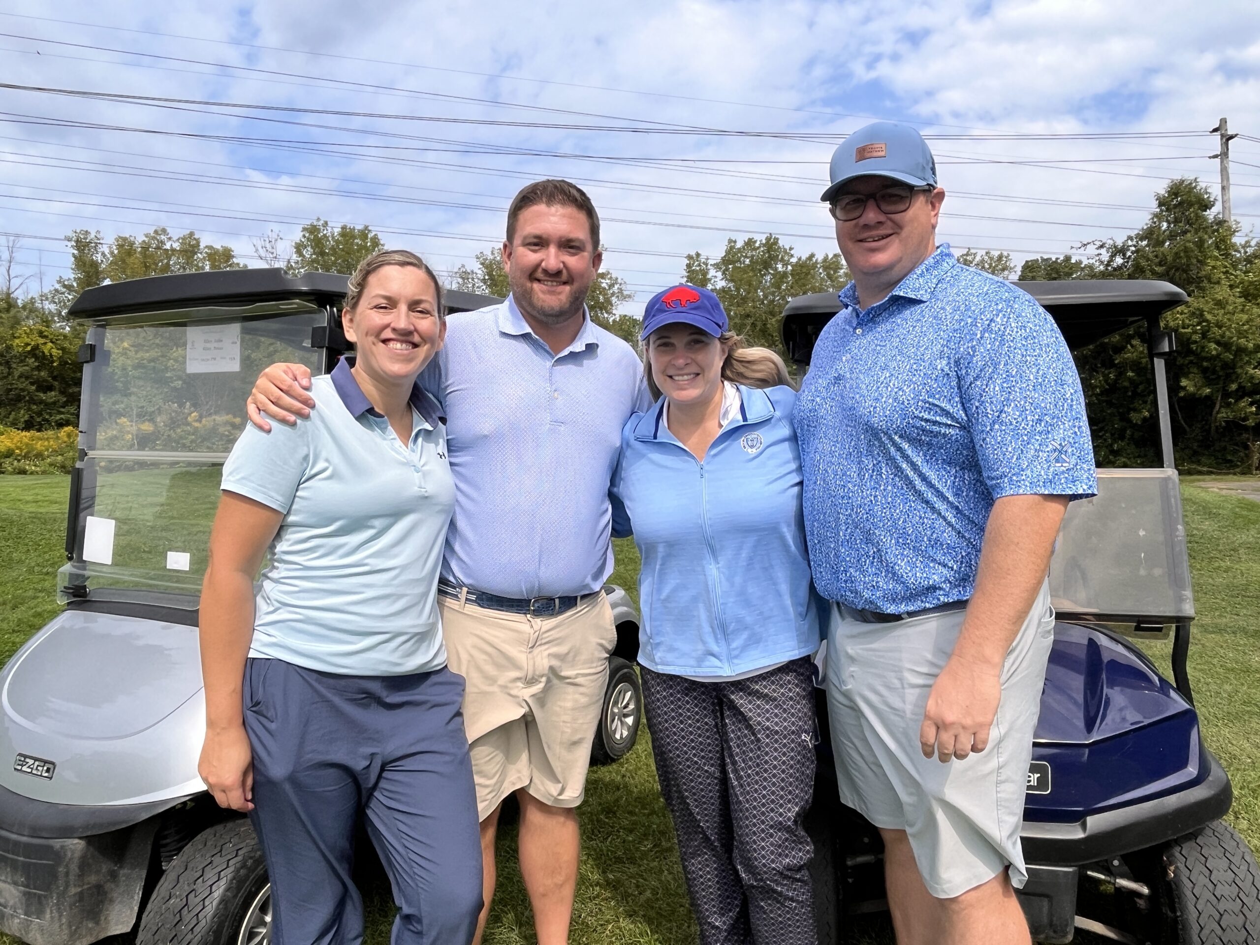two men and two women stand smiling in front of two golf carts on a golf course