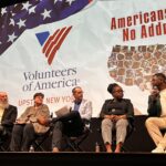 Five people sit on stage in front of a screen with VOA Upstate New York logo as they discuss the "Americans with No Address" documentary