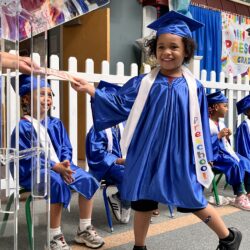 Preschool age girl in blue graduation robes accepts certificate with a smile.