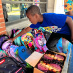 Young kid reaches for a backpack in a pile of backpacks.