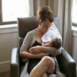 Woman sits in chair cradling newborn baby.
